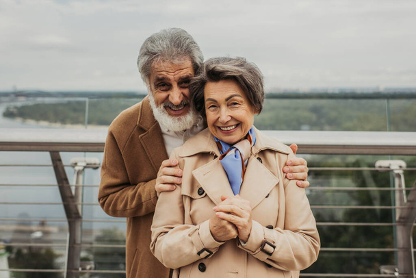 cheerful man with beard hugging senior wife in trench coat outdoors  - Fotó, kép
