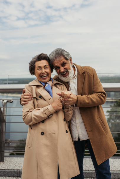 fröhlicher Mann mit Bart umarmt Seniorin im Trenchcoat draußen - Foto, Bild