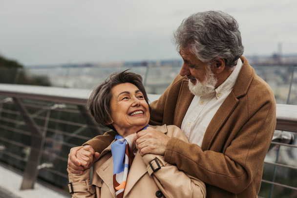 heureux senior homme étreignant femme positive en manteau beige et souriant à l'extérieur  - Photo, image