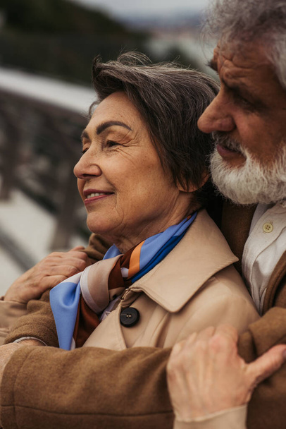 portrait of bearded senior man hugging positive wife in beige coat and smiling outside  - Fotó, kép