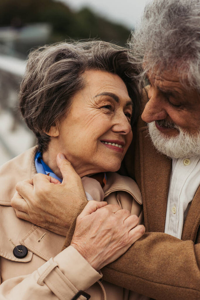 portrait of happy senior man hugging pleased wife in beige coat and smiling outside  - Fotó, kép
