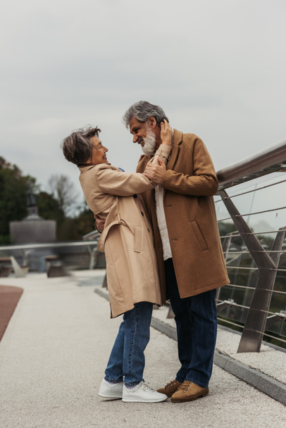 in voller Länge glückliche ältere Frau umarmt bärtigen Ehemann in beigem Mantel auf Brücke in der Nähe der Leitplanke  - Foto, Bild