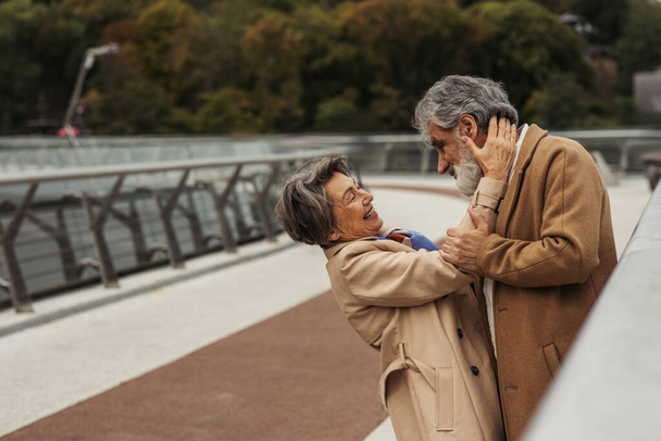 cheerful elderly woman hugging bearded husband in beige coat on bridge near guard rail  - Photo, Image