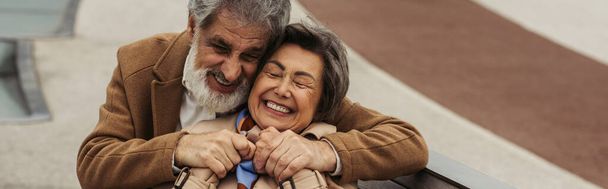 cheerful senior man in coat hugging elderly wife smiling with closed eyes - Foto, afbeelding