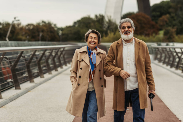 Fröhliche Seniorin spaziert mit glücklichem Mann mit Regenschirm auf Brücke  - Foto, Bild