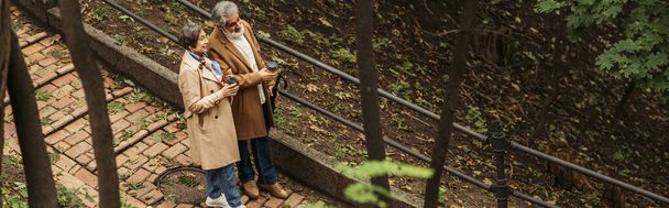 high angle view of cheerful and retired couple in beige coats holding paper cups while walking in park, banner - Φωτογραφία, εικόνα