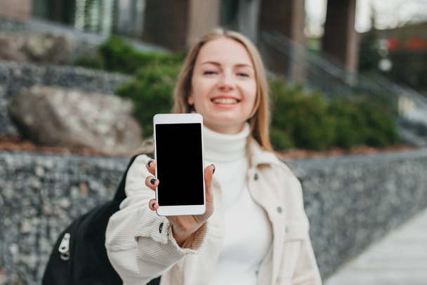 Menina estudante sorrindo feliz mostrando telefone celular contra o fundo da faculdade. Mockup para design - Foto, Imagem