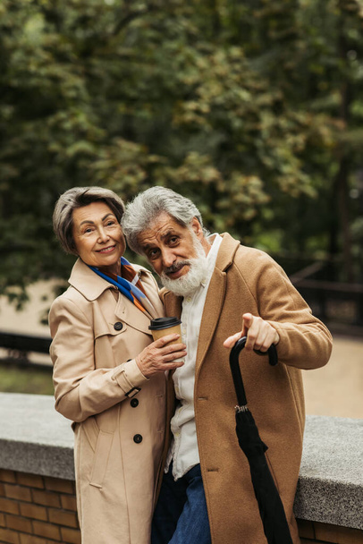 idős férfi kezében esernyő és ujjal mutogatott feleség mellett papír pohár  - Fotó, kép