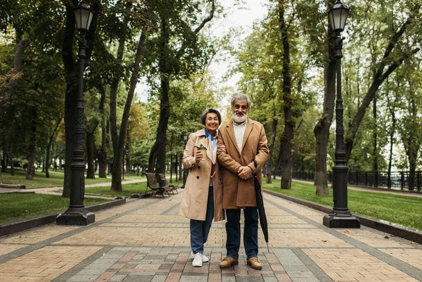 full length of senior woman holding paper cup while standing with stylish husband in coat - Foto, Imagen