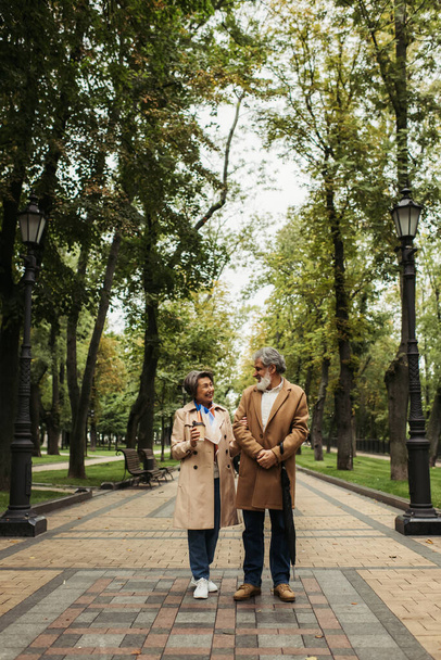 volle Länge der glücklichen Seniorin mit Pappbecher beim Gehen mit stilvollem Mann im Mantel - Foto, Bild