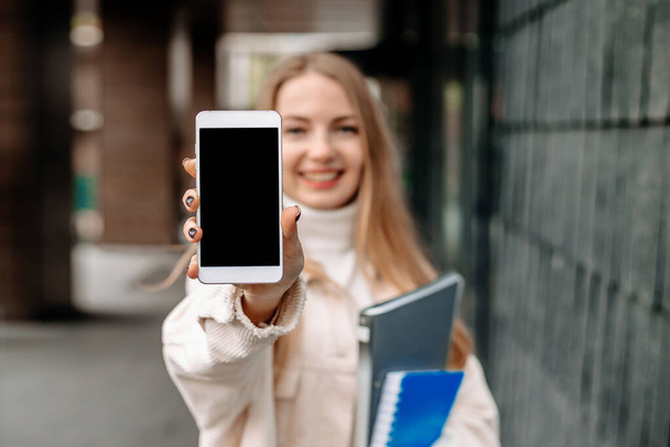 Feliz chica estudiante rubia sonriente mostrando la pantalla de un teléfono móvil en el fondo de la universidad. Burla para el diseño - Foto, imagen