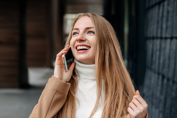 Young blonde Caucasian woman in a beige coat talking on a mobile phone, smiling and looking up against the background of an office building. Corporate employee. Happy female IT-specialist - Photo, Image