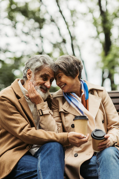 joyful senior couple in coats holding paper cups and sitting on bench in park  - Photo, Image