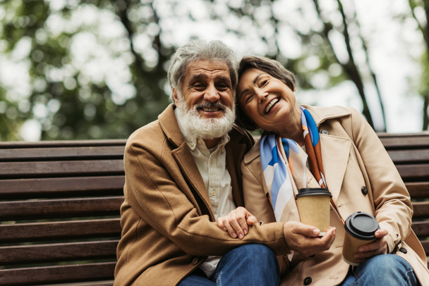 heureux couple âgé en manteaux tenant des tasses en papier avec du café pour aller et assis sur le banc dans le parc  - Photo, image