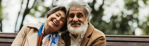 happy senior couple in coats smiling while looking at camera in park, banner - Photo, Image