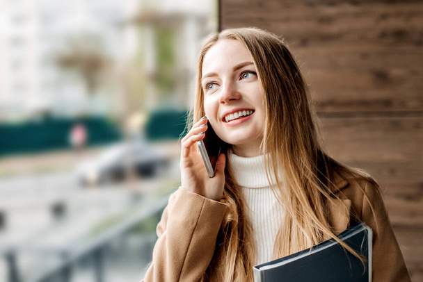 Nuori blondi nainen puhuu matkapuhelimella hymyillen ja katsoen poispäin toimistorakennuksen taustaa vasten. Yhtiön työntekijä. Tietotekniikka-asiantuntija - Valokuva, kuva