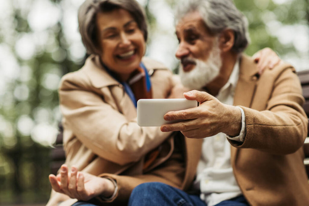 blurred senior woman smiling while bearded husband in coat taking selfie on focused smartphone - Фото, изображение