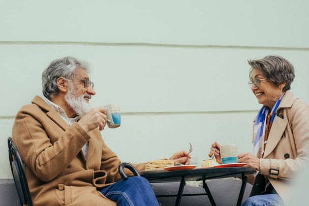 眼鏡とトレンチコートを身に着けた明るい先輩女性が幸せな夫とカフェのテラスでブランチ - 写真・画像