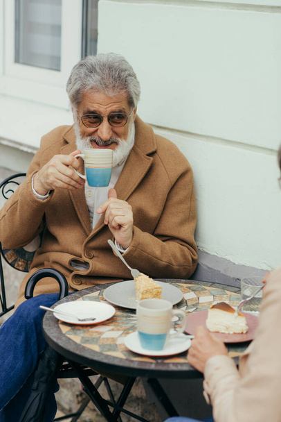 bearded senior man in coat and eyeglasses drinking tea near cakes and wife on terrace of cafe  - Фото, изображение