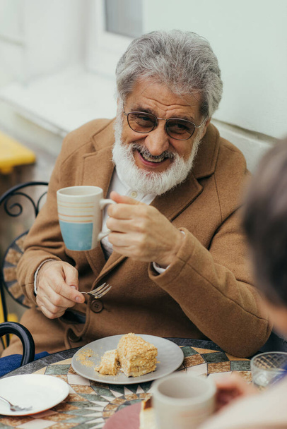 весёлый пожилой мужчина в пальто и очках держит чашку чая возле торта и жену на террасе кафе  - Фото, изображение
