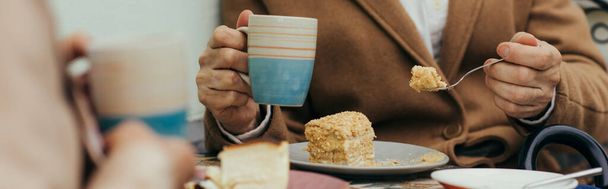 cropped view of senior man in coat holding cup of tea and eating cake near wife on terrace of cafe, banner - Foto, Imagem