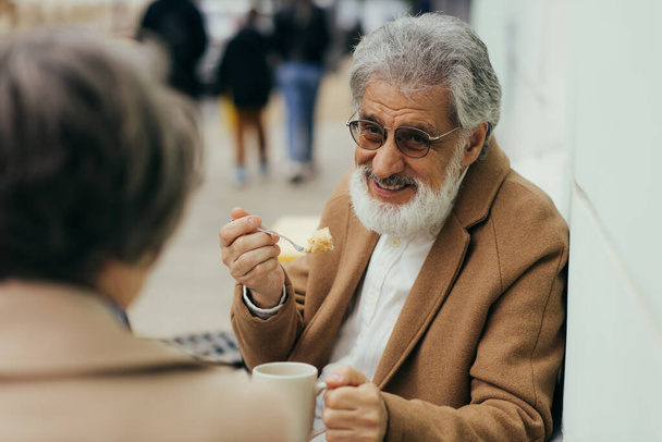 gelukkig senior man in jas houden kopje thee en het eten van taart in de buurt van vrouw op terras van cafe  - Foto, afbeelding