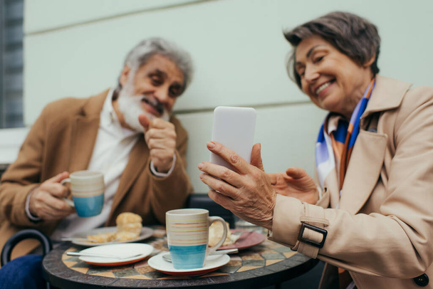 счастливая пожилая женщина держит смартфон во время завтрака с мужем на террасе кафе - Фото, изображение