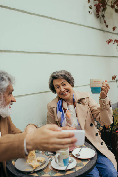bearded senior man taking selfie with cheerful wife during breakfast on terrace of cafe  - Zdjęcie, obraz
