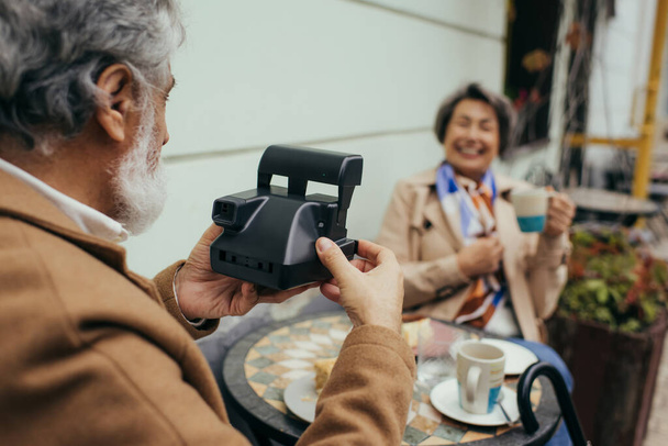 man met baard houden vintage camera en het nemen van foto van wazig vrouw tijdens het ontbijt  - Foto, afbeelding