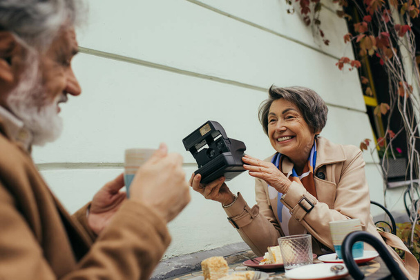 glückliche Seniorin hält Vintage-Kamera neben verschwommenem Mann mit Tasse Tee - Foto, Bild