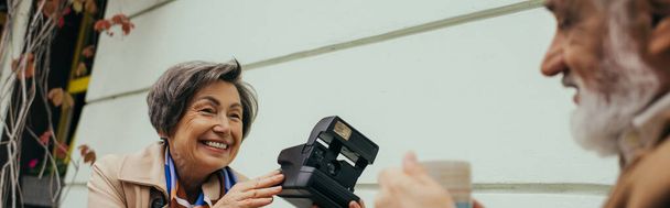 happy senior woman holding vintage camera near blurred husband with cup of tea, banner - Φωτογραφία, εικόνα