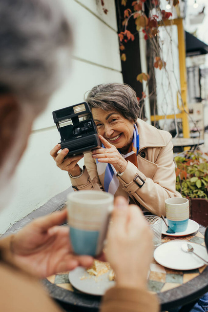 feliz mujer mayor sosteniendo la cámara vintage y tomando la foto de marido borroso con taza de té - Foto, Imagen