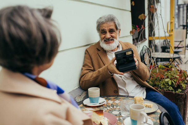 felice uomo anziano con la barba tenendo fotocamera vintage durante la colazione con la moglie  - Foto, immagini