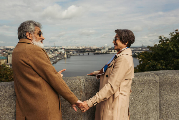 pareja jubilada en abrigos beige tomados de la mano y mirándose el uno al otro cerca del río con vista a la ciudad  - Foto, imagen