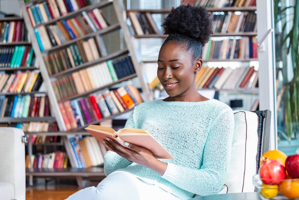 Νεαρή όμορφη Αφρο-Αμερικανίδα διαβάζει ένα βιβλίο στον καναπέ με τα ράφια της βιβλιοθήκης στο πίσω μέρος. Όμορφη γυναίκα σε ένα λευκό καναπέ διαβάζοντας ένα βιβλίο - Φωτογραφία, εικόνα