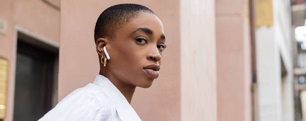 Trendy african american woman with short hair listening music in earphone on street in Treviso, banner  - Foto, afbeelding