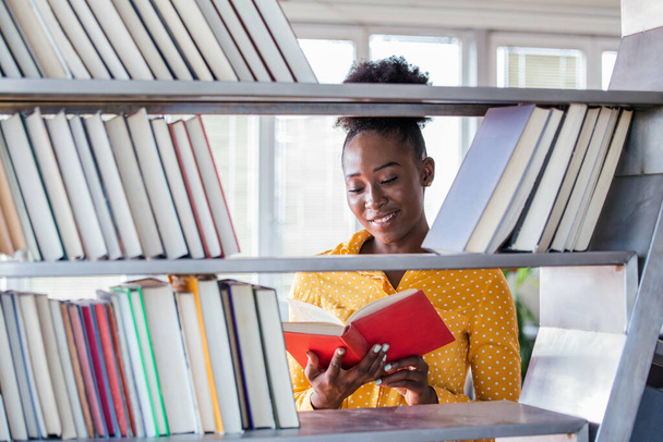 Een Afro-Amerikaanse studente die in de bibliotheek werkt met een boek in de hand, ziet er slim uit. boekenplanken in de bibliotheek. - Foto, afbeelding