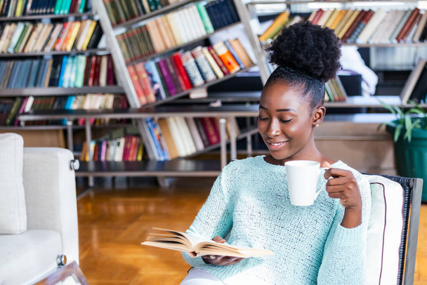 Garota afro-americana lendo um livro sorrindo feliz e relaxado em um dia de lazer em casa. Biblioteca em casa com estantes na parte de trás. - Foto, Imagem