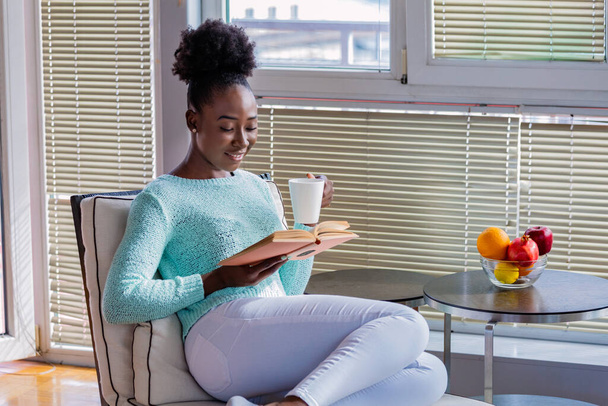 Jolie jolie jeune femme lisant le livre et buvant du café sur le canapé assis près des fenêtres. photo de femme sur le canapé avec vieux livre et tasse de café - Photo, image