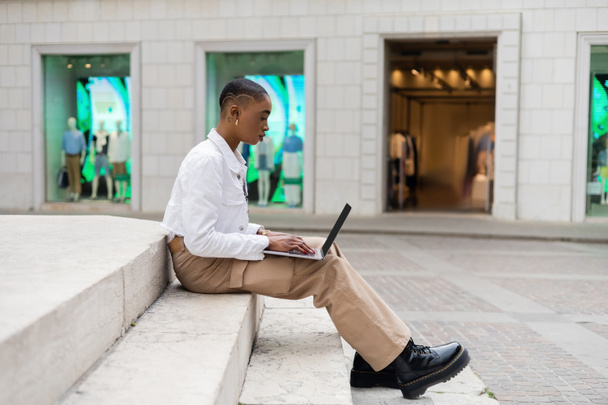 Side view of stylish african american freelancer using laptop with blank screen on stairs on urban street in Italy - Photo, Image