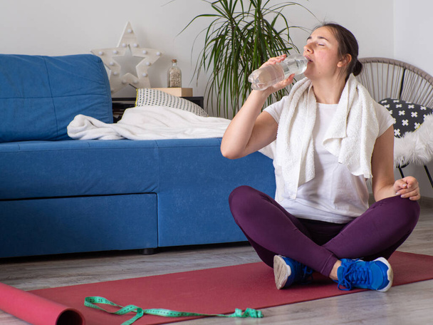 European girl drinking water after practising yoga. Concept of fitness, sport at home and healthy lifestyle. Online training, watching video tutorial on a laptop. Woman sitting on yoga mat. - Zdjęcie, obraz