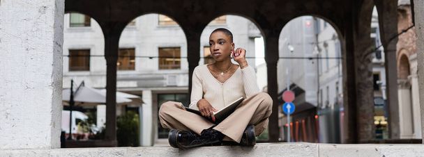 Mujer afroamericana de pelo corto de moda sosteniendo cuaderno al aire libre en Treviso, pancarta - Foto, Imagen