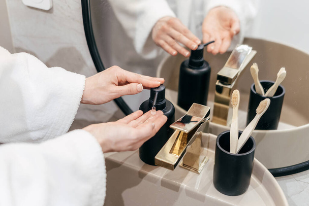 Cropped view of woman dressed in white bathrobe washing hands in the bathroom at home, using antibacterial soap in dispenser bottle. Concepts of hygienic procedures - Photo, Image