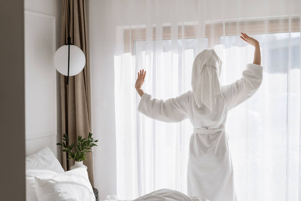 Vrouw in badjas die 's morgens wakker wordt, staat bij het raam in de hotelkamer, strekt zich uit, steekt haar handen op. Vrouwelijke rust na douche thuis of in het kuuroord. Comfortabel appartement, recreatie concept - Foto, afbeelding