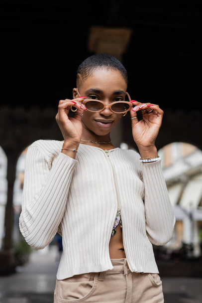 Fashionable african american woman holding sunglasses and looking at camera on urban street  - Valokuva, kuva