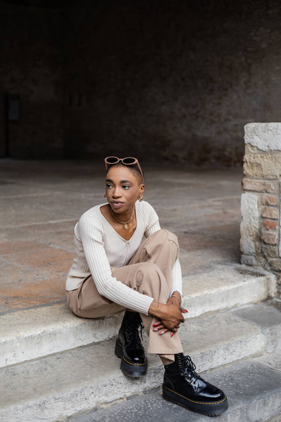 Trendy african american woman with sunglasses sitting on stairs on urban street in Treviso - Foto, Imagen
