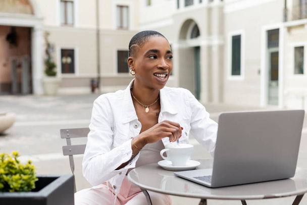 Positive african american freelancer looking away near coffee and laptop in outdoor cafe in Italy - 写真・画像