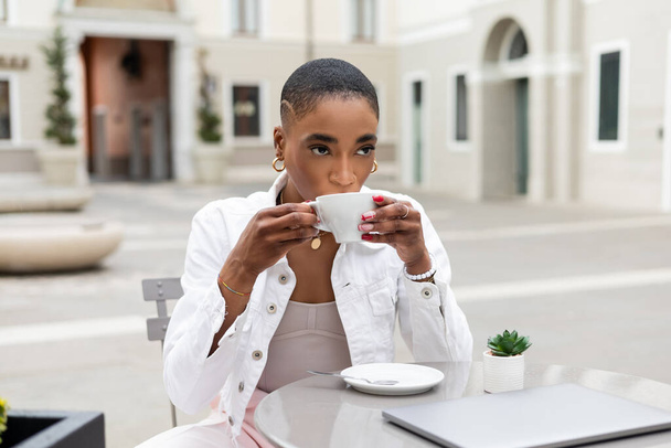 イタリアの屋外カフェでラップトップに近いスタイリッシュなアフリカ系アメリカ人ブロガーコーヒーを飲む - 写真・画像