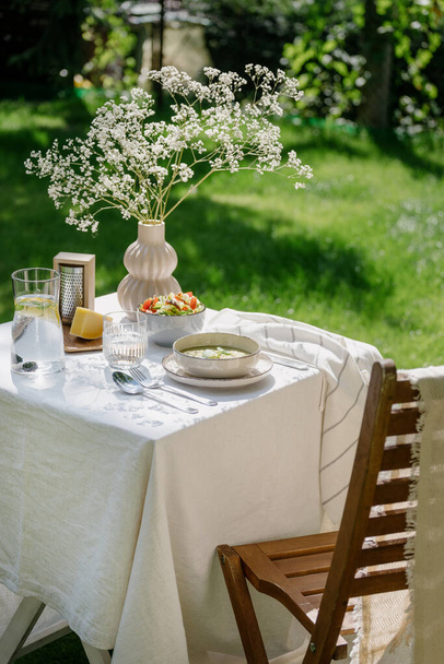 houten stoel en tafel serveert wit linnen tafelkleed, verse groentesalade, kaas, soep in kom, koud water met citroen in glazen kan en bloemen in vaas in het park buiten in de zomer - Foto, afbeelding