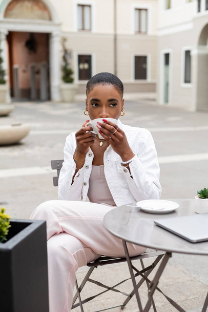 Fashionable african american woman drinking coffee near laptop in outdoor cafe in Italy - Photo, Image